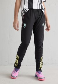 Melni sporta bikses ar rozā akcentiem un dzeltenām svītrām. Satur Juventus un Adidas logo. Valkātas ar krāsainām, rakstainām sporta apaviem.