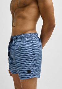Swim shorts in blue with a textured fish scale pattern, elastic waistband with drawstring, and small black logo patch on the side.