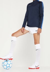 Osoba w granatowej i niebieskiej długiej koszulce sportowej, białych szortach i skarpetkach, balansująca lewą stopą na białej i niebieskiej piłce nożnej.