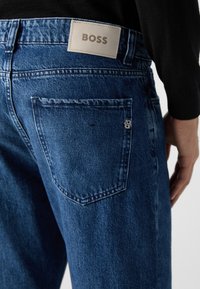 Pantalones vaqueros de mezclilla azul con un corte regular, que presentan una etiqueta de cuero etiquetada como "BOSS" en la cintura y un acento en el bolsillo trasero.