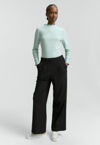 Kobieta stoi z rękami w kieszeniach, ubrana w jasnoniebieską bluzkę z długim rękawem, czarne szerokie spodnie i białe sneakersy na jednolitym tle.