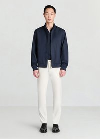 Veste zippée bleu marine en tissu lisse, avec poches latérales et col montant, associée à un pantalon blanc et des chaussures en cuir noir.