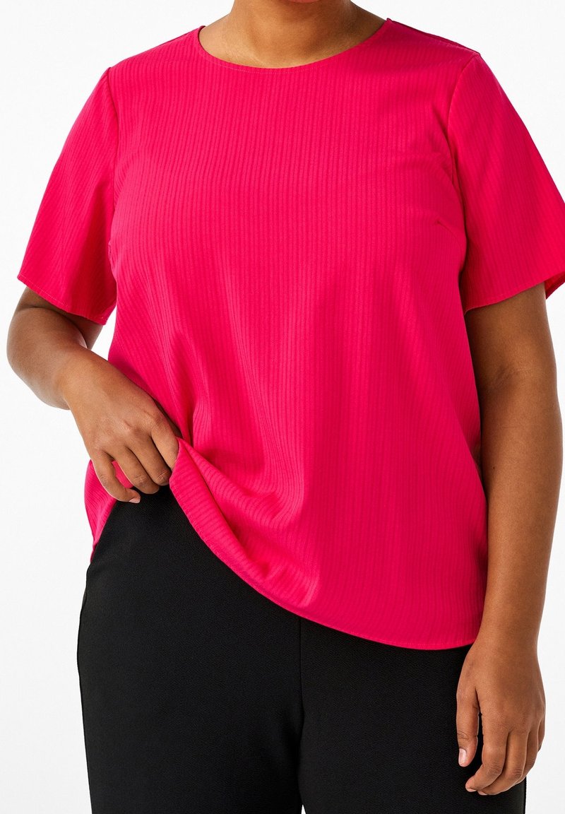 Femme portant un chemisier rose vif à manches courtes et un pantalon noir, tenant l'ourlet du chemisier avec la main gauche.