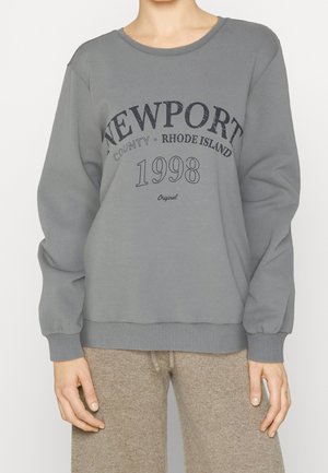 Osoba nosząca szarą bluzę z napisem "NEWPORT County Rhode Island 1998 Original" i beżowe spodnie na jednolitym tle.