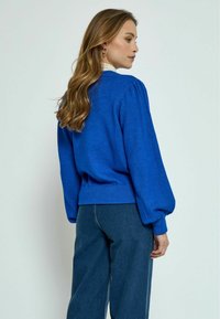 Modra pulover s puffastimi rokavi in ribastim robom. Mehak material, v kombinaciji s temno jeans hlačami. Model je prikazan s hrbtne strani.
