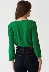 Bluză verde cu mâneci lungi și croială lejeră, având un decolteu larg și ușoare pliuri la umeri, asortată cu blugi de culoare albastru închis.