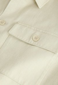 Gros plan sur un tissu beige montrant une poche à rabat boutonnée et une partie du col d'un vêtement.