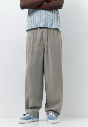 Személy szürke, bőszárú nadrágot, világoskék kábelmintás ujjatlan pulóvert és kék, fűzős sportcipőt visel, karját keresztbe téve áll.