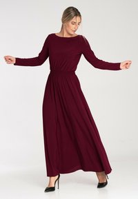 Bordowa sukienka maxi z długim rękawem, zebrana w talii, płynący dół i gładki materiał. Czarne buty na wysokim obcasie dopełniają stylizację.