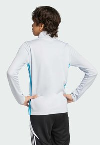 Maglia sportiva bianca a maniche lunghe con collo alto, caratterizzata da strisce laterali blu, design aderente e tessuto liscio. Pantaloni neri visibili.