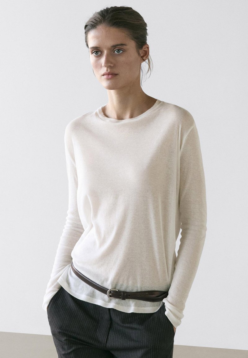 Kvinde med håret samlet bagpå iført en gennemsigtig hvid langærmet top, stoppet ned i mørke bukser med smalle striber og et brunt bælte, hænderne i lommerne.