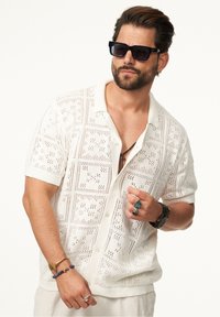Weißes, gestricktes Kurzarmhemd mit strukturiertem, offenem Design. Getragen mit mehreren Ringen und Armbändern, ergänzt durch dunkle Sonnenbrille.