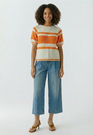 Smiling woman with curly hair wearing an orange and beige striped short-sleeve sweater, wide-leg cropped jeans, and brown heeled sandals.