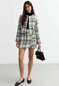 Ensemble composé d'une veste et d'une jupe en tweed dans un motif écossais noir, blanc et gris avec des boutons dorés. Haut noir, ballerines noires et un petit sac à main noir.