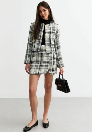 Completo giacca e gonna in tweed con motivo a quadri in bianco, nero e grigio, arricchito da bottoni dorati. Top nero, ballerine nere e una piccola borsa nera.