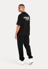 Sort oversized t-shirt med hvid tekst på ryggen, sorte sweatpants og hvide sneakers. Glat stof, afslappet design, løs pasform.