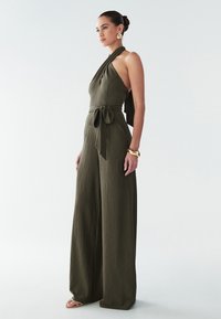Olive green halter jumpsuit with a textured fabric, wide-leg design, and tied waist detail. Model wearing gold earrings and sandals.