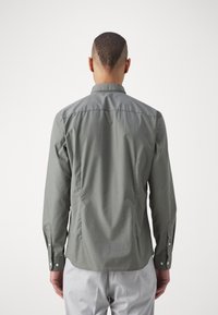 Camisa verde de manga larga con cuello recto, puños abotonados, pliegues en la espalda y una textura suave; llevada sobre pantalones de color claro.