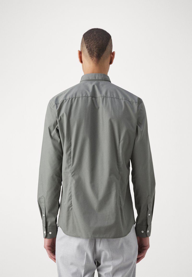 Grünes Langarmhemd mit einem geraden Kragen, knöpfbaren Manschetten, Rückenfalten und glatter Textur; getragen über hellen Hosen.