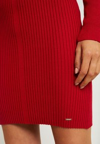 Rotes, geripptes Strickkleid mit taillierter Passform, lange Ärmel und einem metallischen Markenetikett nahe dem Saum. Glatte Textur und elastische Passform.