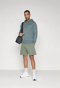 Camisola com capuz cinzento-azulada com bolso canguru, combinada com calções verde claros. Ténis brancos e mala preta tiracolo. Corte justo. Design simples.