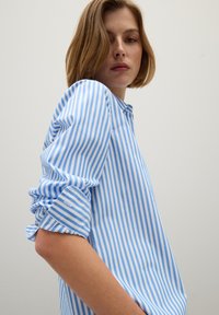 Chemise à rayures bleues et blanches à manches longues avec patte de boutonnage, manches froncées et tissu en coton léger. Texture lisse et coupe décontractée.
