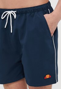 Marineblå badebukser med hvit snor, sidelommer og brodert logo med røde og gule detaljer på kanten.