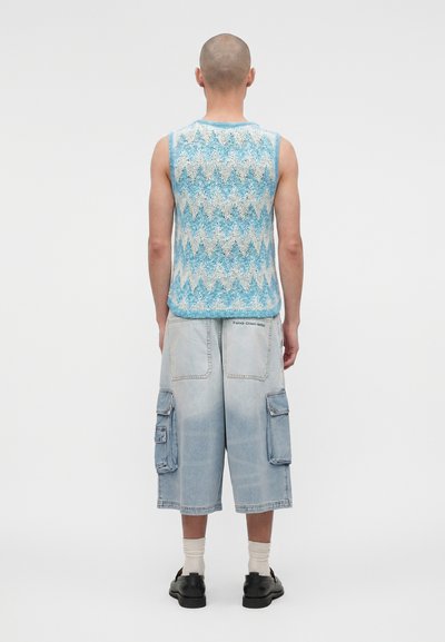 Άνδρας με ξυρισμένο κεφάλι φοράει αμάνικη μπλε και λευκή πλεκτή μπλούζα με ζιγκ-ζαγκ μοτίβο, ανοιχτό μπλε cargo σορτς, λευκές κάλτσες και μαύρα παπούτσια, στέκεται με την πλάτη γυρισμένη.