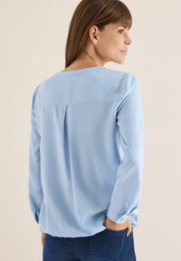 Lys blå langærmet bluse med rund halsudskæring, lægdetalje bagpå og knaplukning ved ærmerne. Glat stofstruktur, afslappet design.