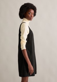 Zwart denimjurk met schouderbandjes, lichte A-lijnvorm. Gedragen over een lichtcrème coltrui met lange mouwen.