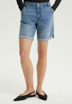 Jasnoniebieskie szorty dżinsowe z podwiniętym brzegiem, noszone z czarnym topem z długim rękawem oraz czarnymi spiczastymi balerinami na paseczku z tyłu. Ręce w kieszeniach.