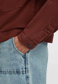 Hand in light blue denim jeans pocket, wearing a textured dark red long sleeve shirt with a buttoned cuff.