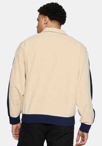 Beżowa kurtka polarowa z granatowymi akcentami. Posiada kołnierz, długie rękawy oraz ściągacze przy mankietach i u dołu. Miękka w dotyku.