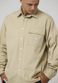 Camicia beige a maniche lunghe in cotone, con taschino sul petto e una targhetta colorata. Bottoni sul davanti e design semplice.