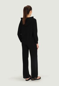 Czarna bluza z kapturem i pasujące szerokie spodnie z miękkiego materiału. Spodnie mają rozcięcia po bokach; noszone z płaskimi sandałami. Minimalistyczny design.