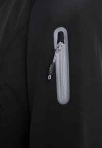Gros plan d'un tissu noir avec une poche zippée verticale grise dotée d'un cordon gris et d'une étiquette noire portant la mention CMP.
