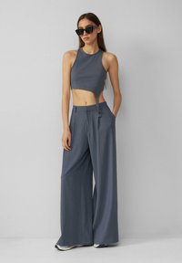 Sivina rebrasta kratka majica s izrezom sa strane, uparena sa širokim sivim hlačama. Model nosi oversize crne naočale i patike.