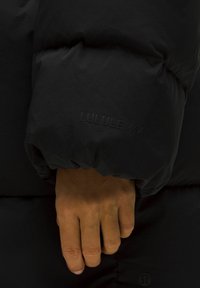 Manica di giacca a vento nera con polsino a coste, caratterizzata da un logo Lululemon in rilievo sul tessuto. Texture liscia e opaca, design leggero.