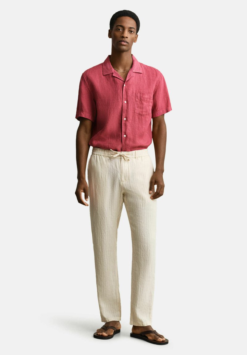 Hombre de pie, vistiendo una camisa roja de manga corta con botones, pantalones crema con cordón y sandalias marrones, contra un fondo blanco liso.