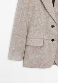 Lysebrun tekstureret blazer med to sorte knapper, en klaplomme foran nederst og en krave med hak, vist fra midtærme til kant.