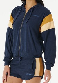 Sweat à fermeture éclair marine avec des rayures dorées et crème, présentant des poignets côtelés, un ourlet avec cordon de serrage et le logo "RIPCURL" sur la poitrine gauche. Associé à un short.