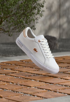 Fehér Lacoste sportcipő bézs részletekkel és logóval, mintázott fa csempék fölött lebegve egy kültéri környezetben, homályos zöld növény és fal közelében.