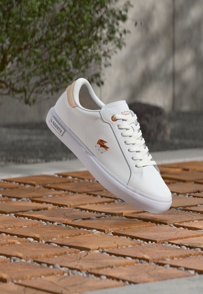 Λευκό αθλητικό παπούτσι Lacoste με μπεζ λεπτομέρειες και λογότυπο, που αιωρείται πάνω από ξύλινα πλακάκια με σχέδια σε εξωτερικό χώρο κοντά σε ένα θαμπό πράσινο φυτό και τοίχο.