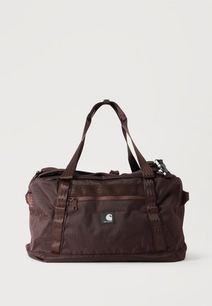 Tmavohnedá taška Carhartt duffel s dvojitými rukoväťami a predným vreckom so zipsom na jednoduchom svetlom pozadí.