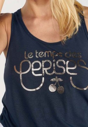 Kvinde iført en mørkeblå ærmeløs top med metallisk guld fransk tekst "Le temps des cerises" og en kirsebærgrafik under teksten.