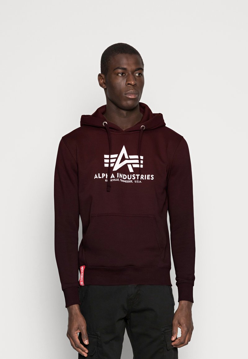 Burgunderfarget hettegenser med en hvit "Alpha Industries" logo og design. Har en lomme foran, snor hette og ribbestrikkede mansjetter.