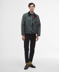 Casaco impermeável verde com fecho de zip, gola preta, detalhe vermelho e bolso frontal. Combinado com jeans escuros, bainhas enroladas e mocassins escuros.