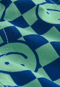 Zöld pamutzsák kék kockás mintával, stilizált mosolygós arcokkal, sima textúrával és steppelt élekkel.