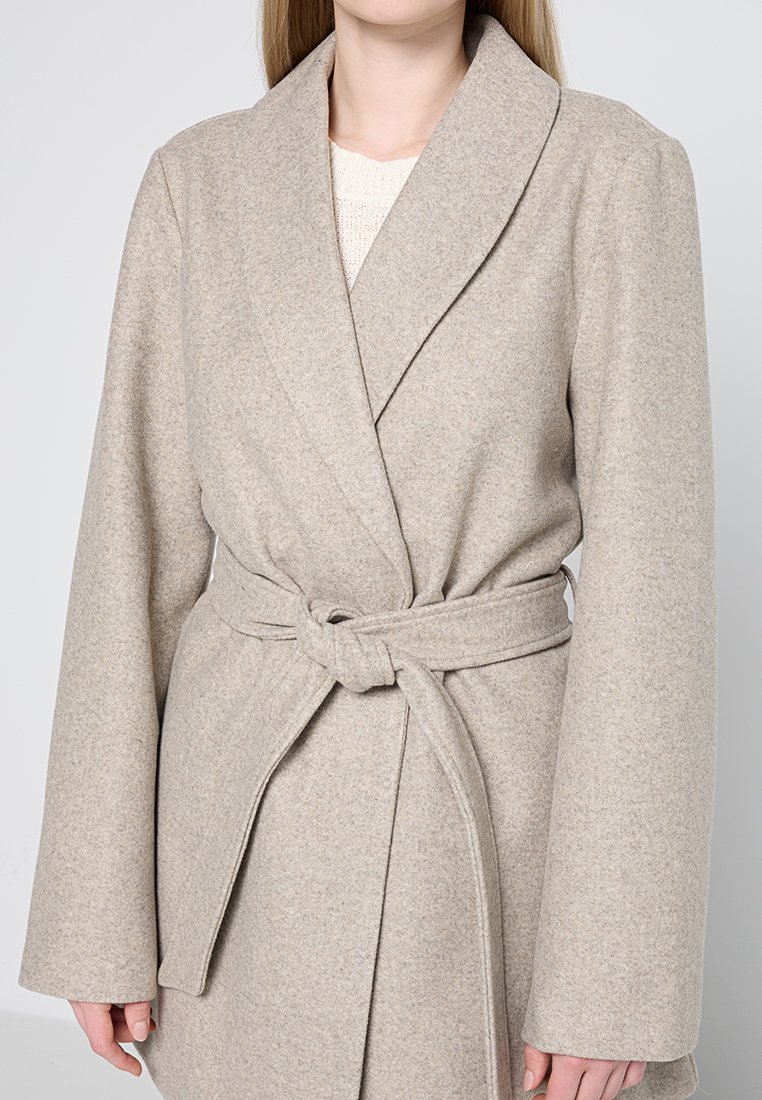 Abrigo envolvente beige hecho de un tejido suave y texturizado. Presenta un cuello ancho, mangas largas y un cinturón anudado en la cintura para cerrarlo.
