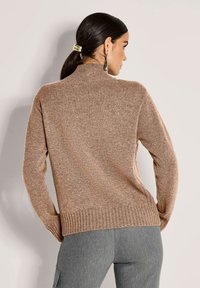 Frau mit dunklem Haar in einem Pferdeschwanz, die eine goldene Haarspange, einen braunen Strickpullover, lange Ohrringe und graue Hose trägt und einer hellen Wand zugewandt ist.
